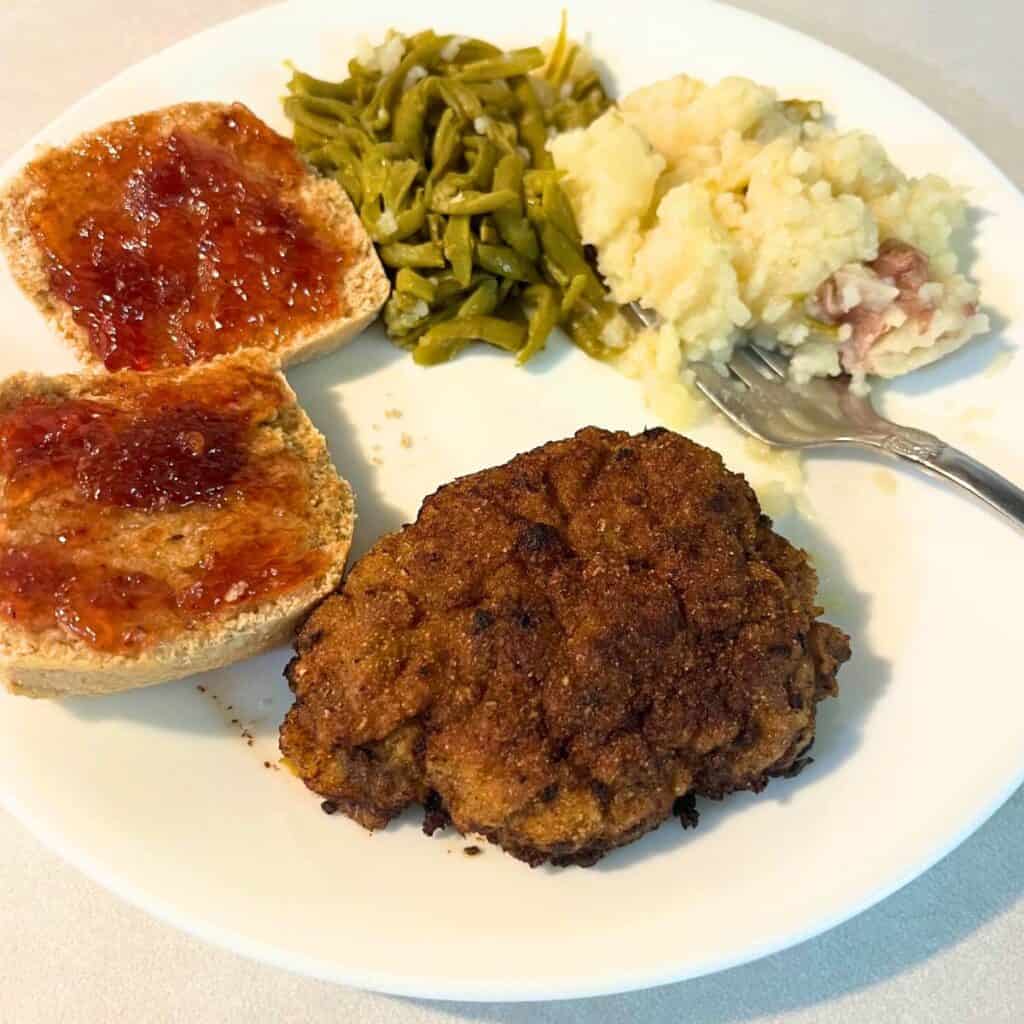 A crispy country fried steak patty, mashed potatoes, green beans, and a biscuit with strawberry preserves on a white plate.