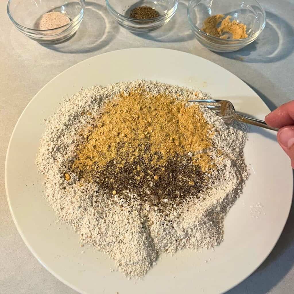 Woman mixing flour, salt, pepper, and seasoned salt on a white plate with a fork.
