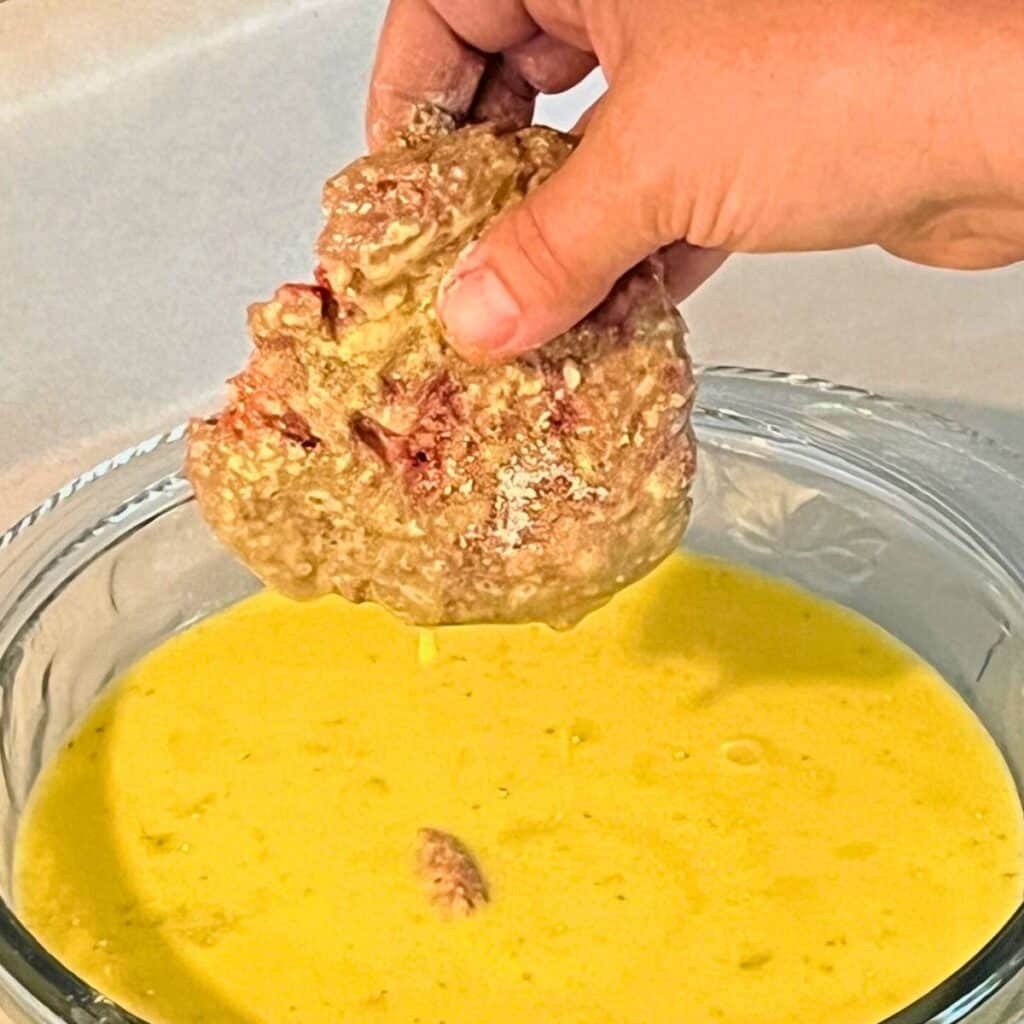 Woman putting a floured beef patty into a bowl of milk and eggs to dredge both sides.