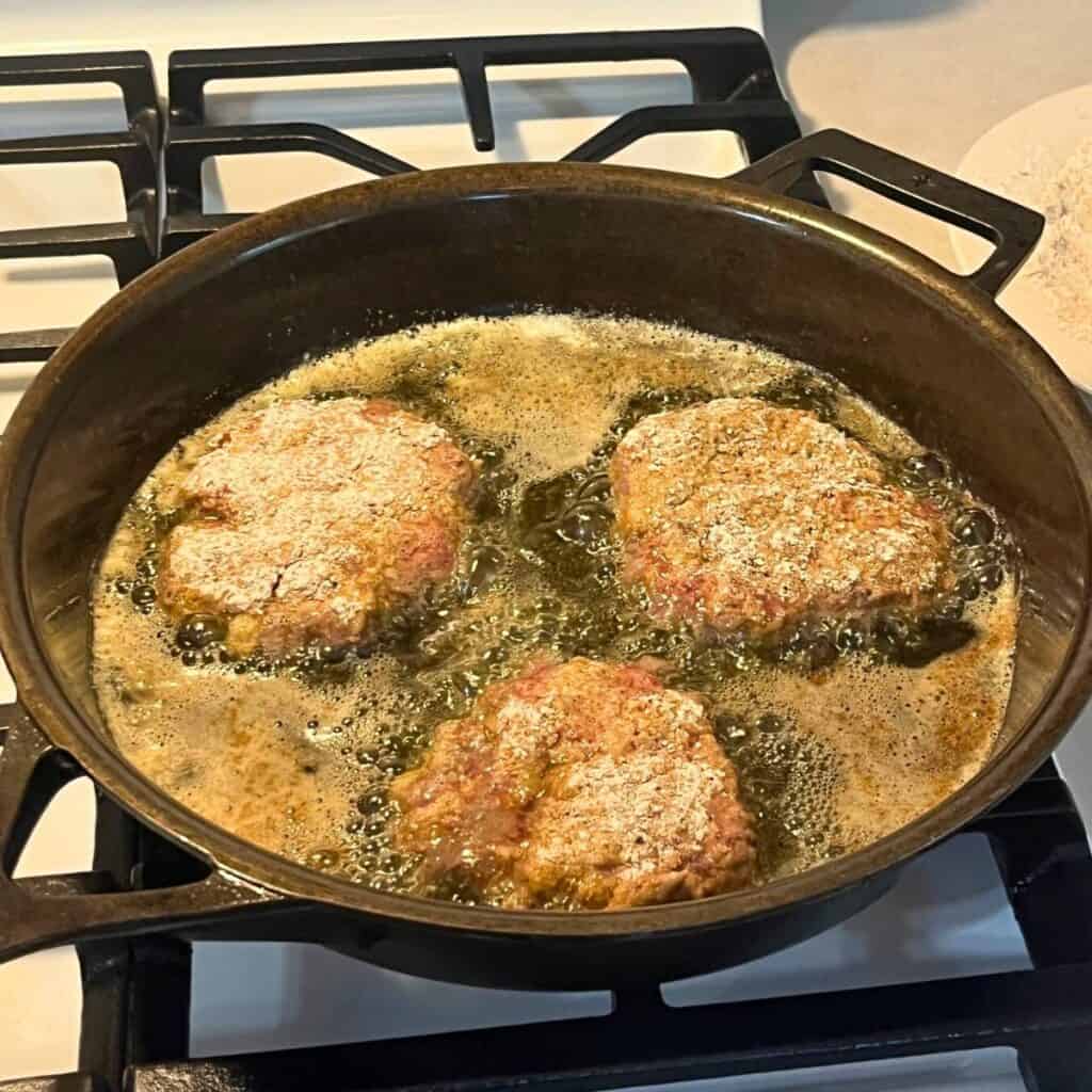Three country fried steak patties in a cast iron skillet on a gas stove.