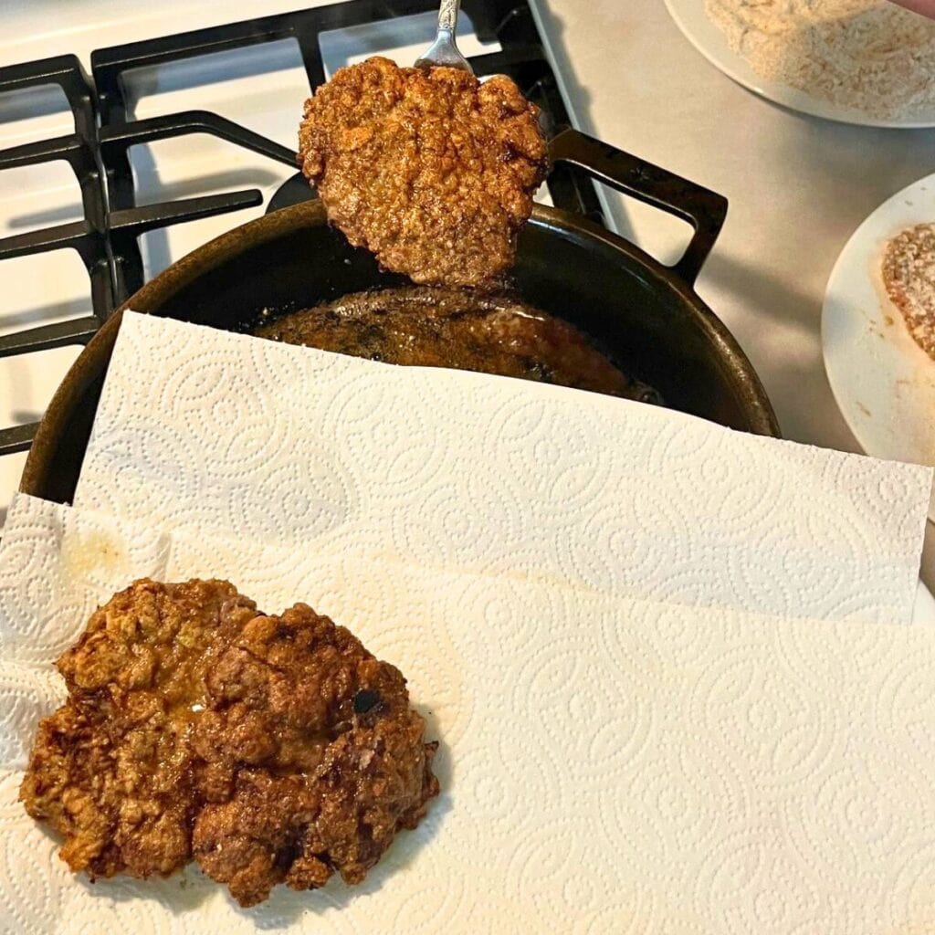 Woman pulling country fried steaks out of a hot cast iron skillet and setting them on a paper towel lined platter to drain.