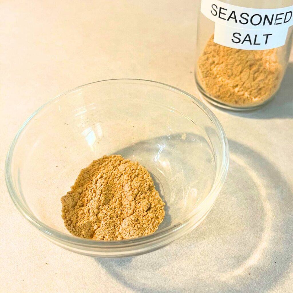 A bowl of homemade seasoned salt sitting on a kitchen counter next to a small glass jar of seasoned salt.