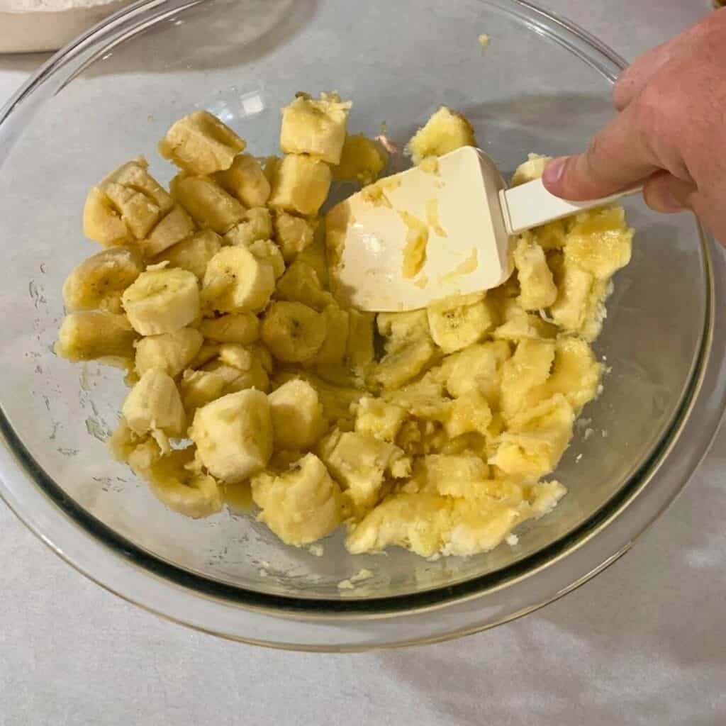 Woman mashing three ripened bananas in a glass mixing bowl with a white rubber spatula.