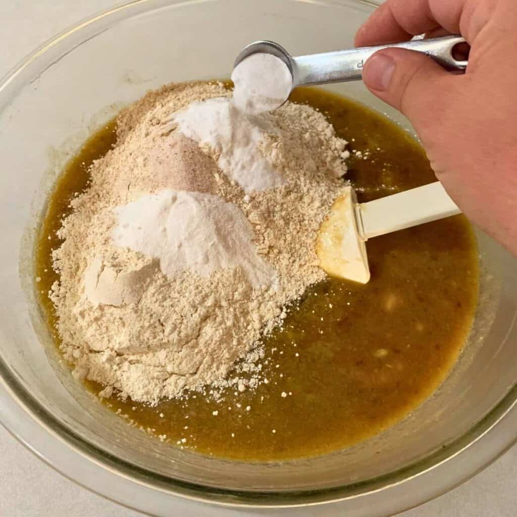 Woman adding dry ingredients to a glass mixing bowl of mashed banana and other wet ingredients for making chocolate banana bread.