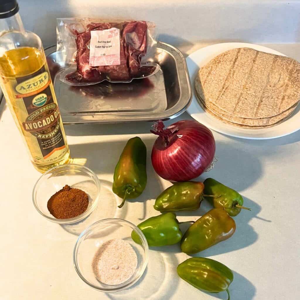 Ingredients to make one pan steak fajitas. Shown are banana peppers, an onion, flour tortillas, salt, chili powder, avocado oil, and beef stew meat.