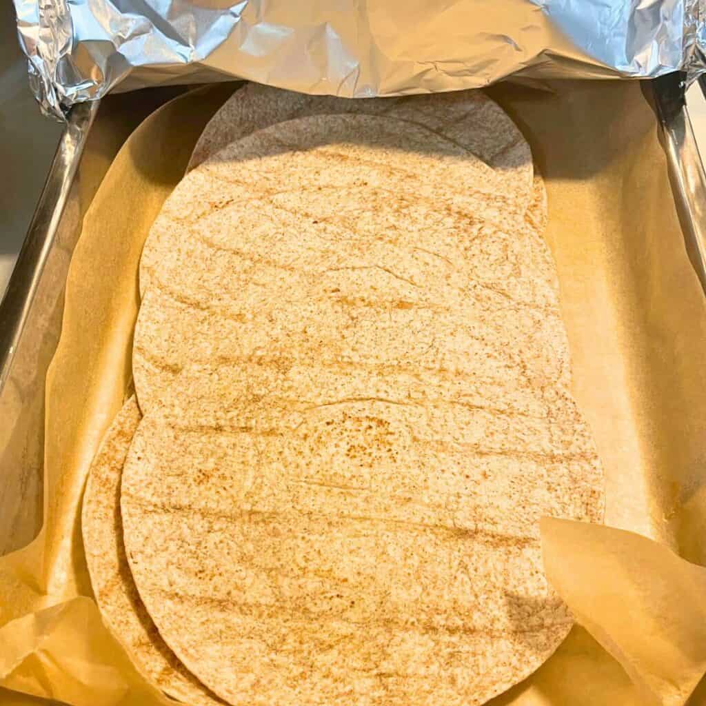 A parchment-lined baking tray of cooked flour tortillas with a foil lid partially removed.