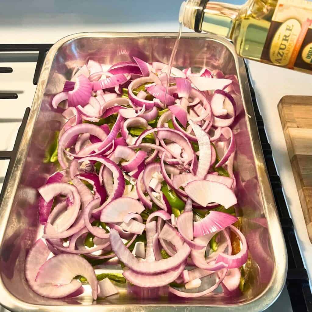 Woman drizzling avocado oil over onion and pepper slices in the bottom of a stainless steel baking pan.