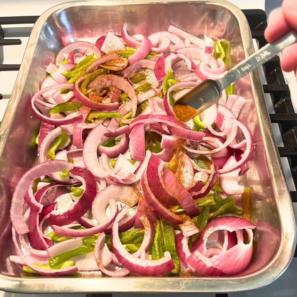 Woman sprinkling chili powder over sliced onions and peppers in the bottom of a stainless steel baking pan.