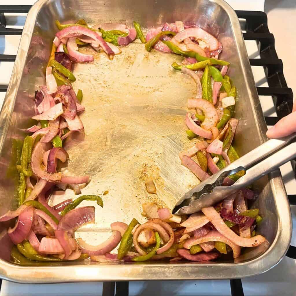 Woman sliding broiled vegetables to the sides of a baking pan with stainless steel tongs.