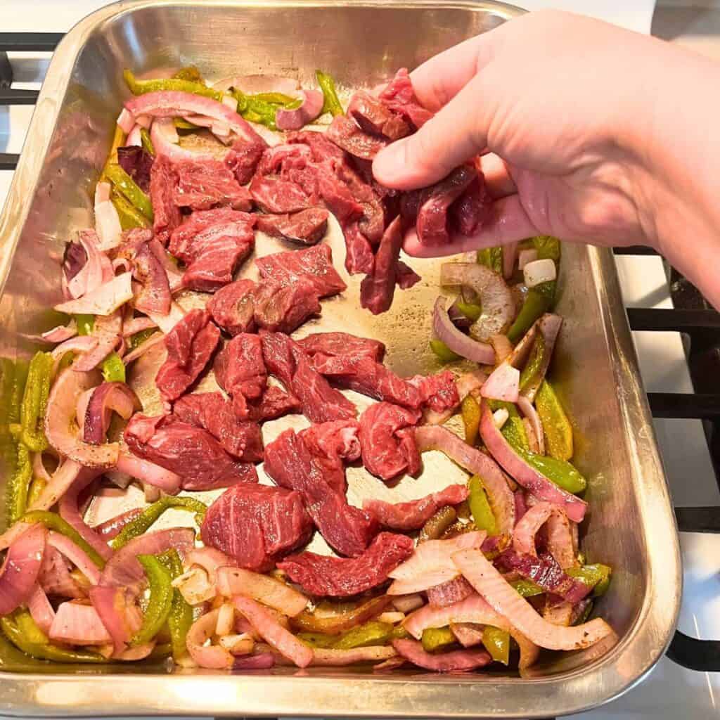 Woman adding sliced beef to the bottom of a baking pan surrounded by broiled onions and peppers.
