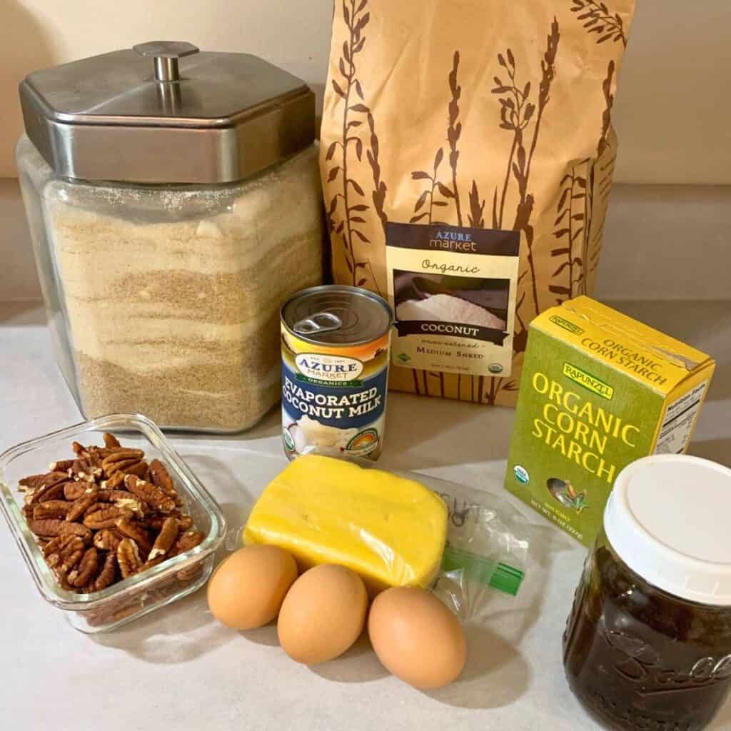 Ingredients for making German chocolate cake icing sitting on a kitchen counter. Pictured is sugar, cornstarch, coconut flakes, coconut milk, pecans, butter, 3 eggs, and vanilla extract.