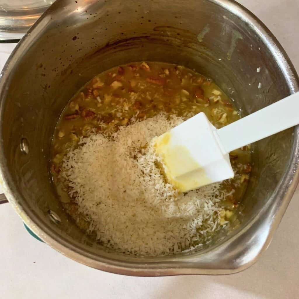 Woman stirring coconut flakes and chopped pecans into a medium saucepan with an egg mixture.