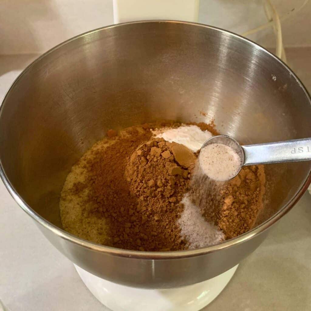 Woman adding a teaspoon of salt into a mixing bowl with flour, sugar, cocoa powder, baking soda, and baking powder.