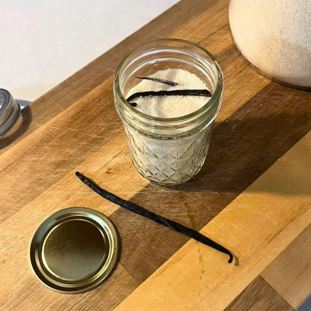 A glass mason jar with vanilla sugar inside sitting on a wood cutting board next to a jar of sugar, vanilla bean, and lid.