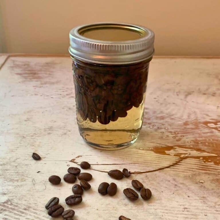 A small mason jar of coffee extract sitting on a white chipped table with coffee beans spilled out.