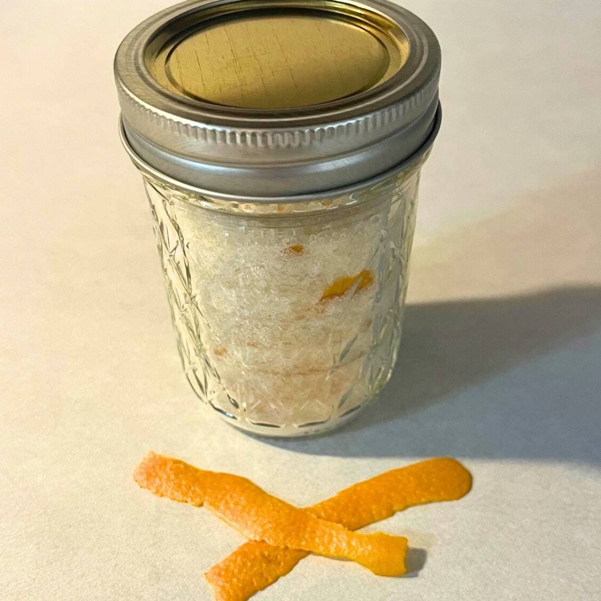 A glass mason jar of orange sugar sitting on a kitchen counter next to wide shreds of orange zest.