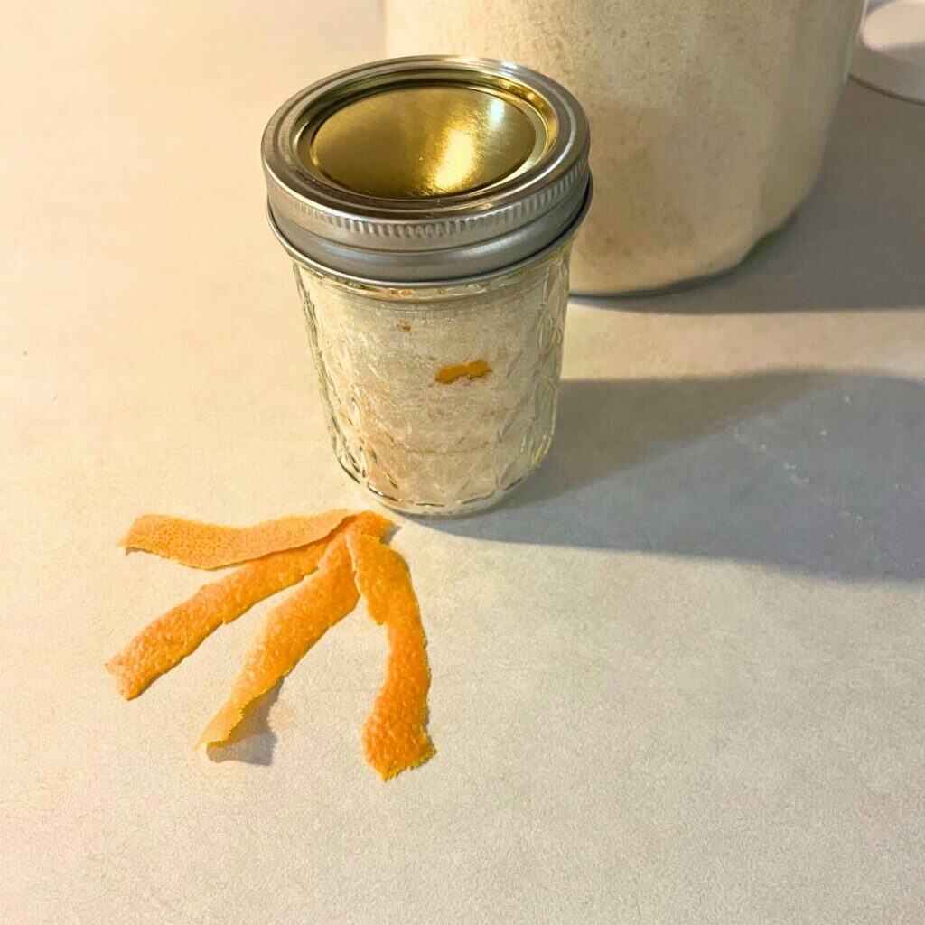A glass jar of orange sugar sitting on a kitchen counter next to another glass jar of sugar and wide strips of orange zest.