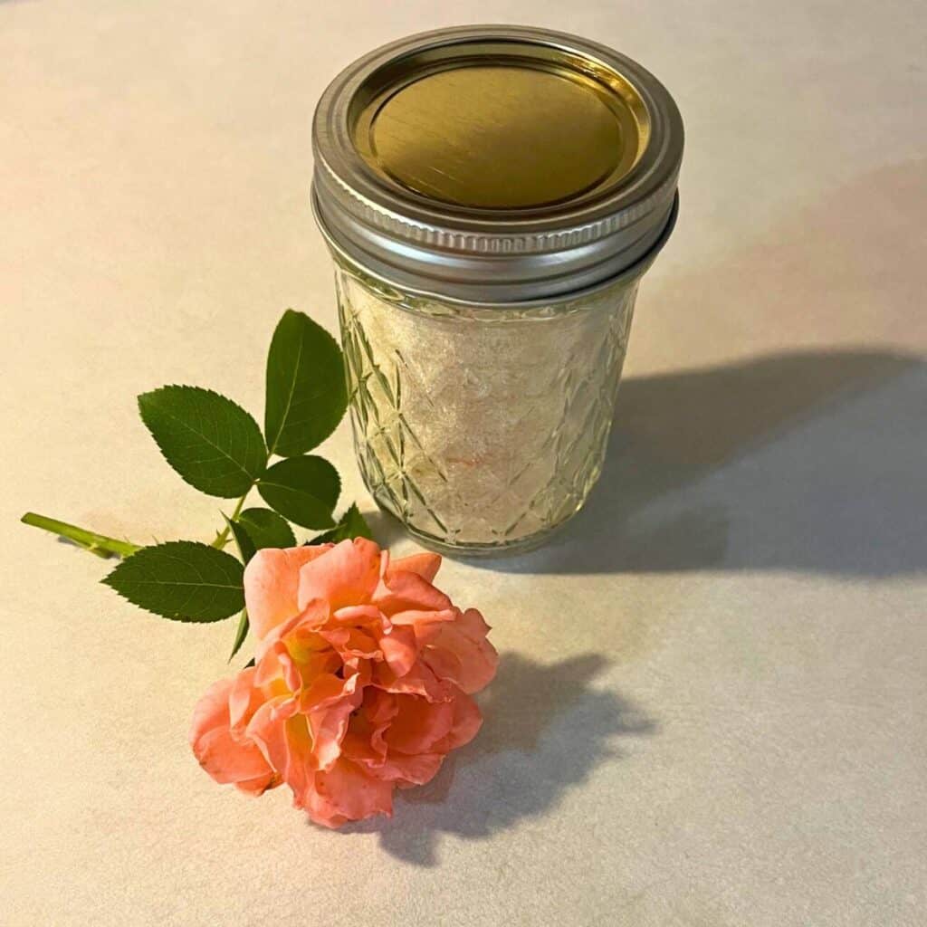 A quilted glass mason jar of rose sugar sitting next to an apricot rose on a kitchen counter.