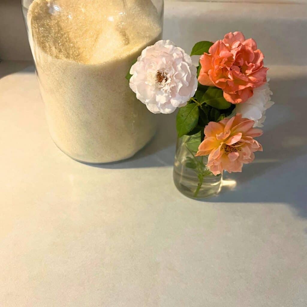 A vase of white, apricot, and pink roses sitting next to a large glass jar of sugar on a kitchen counter.
