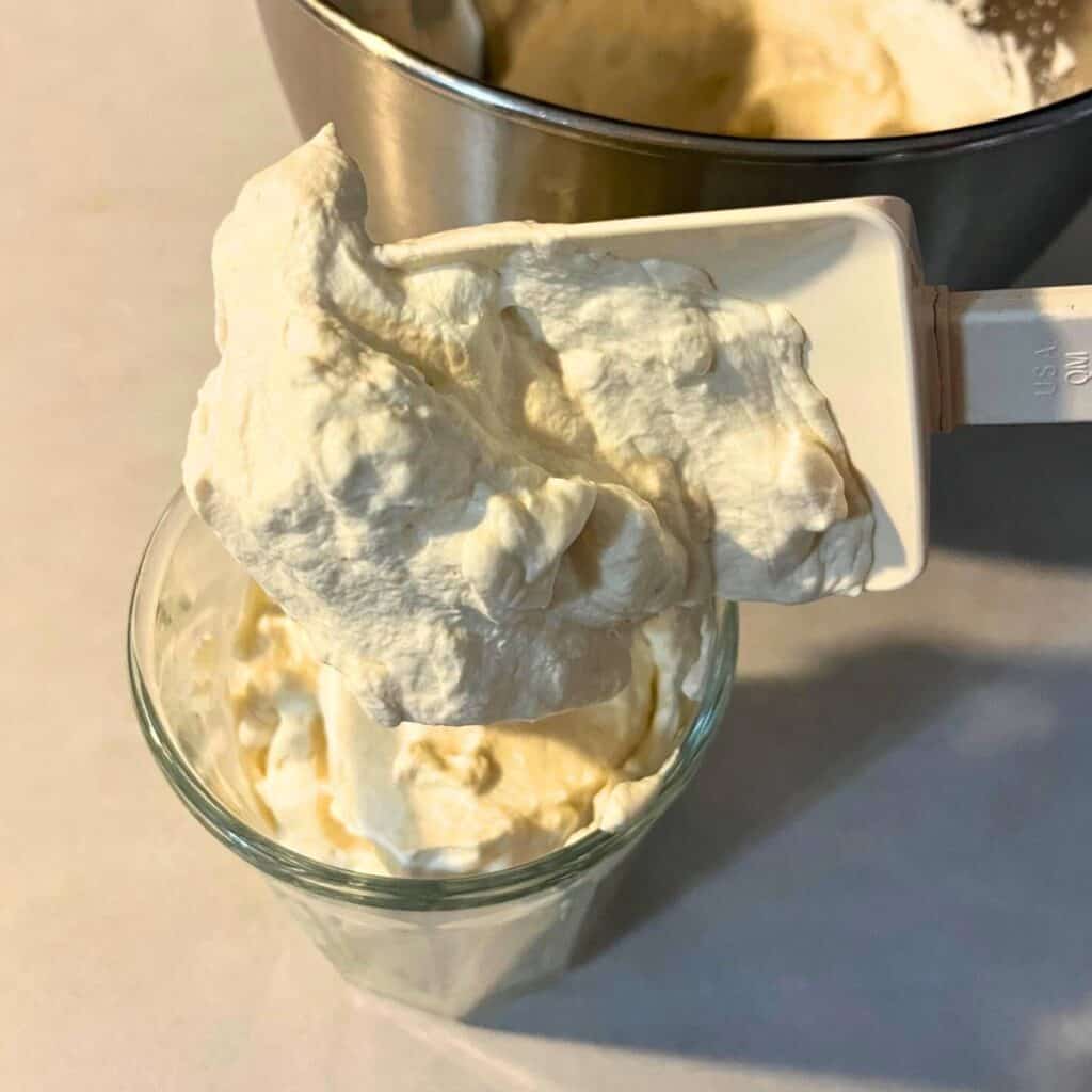 A woman scooping whipped cream out of a stainless steel bowl and putting it into a glass jar with a white rubber spatula.