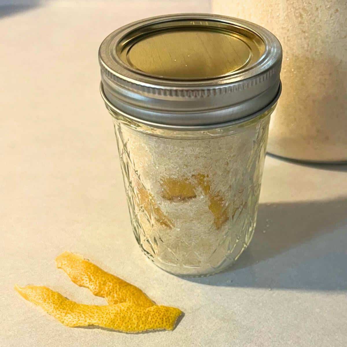 A glass mason jar of lemon sugar sitting on a counter next to strips of lemon zest.