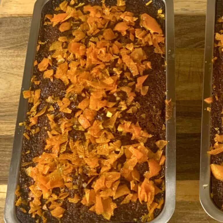 A moist fruit cake made without alcohol sitting in a stainless steel loaf pan on a wood cutting board. The fruit cake has an orange rind topping.