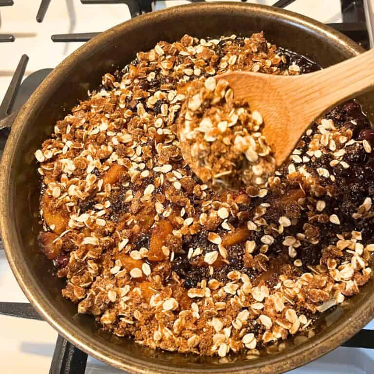 Woman scooping blueberry peach pie from a cast iron skillet on a white gas stove.