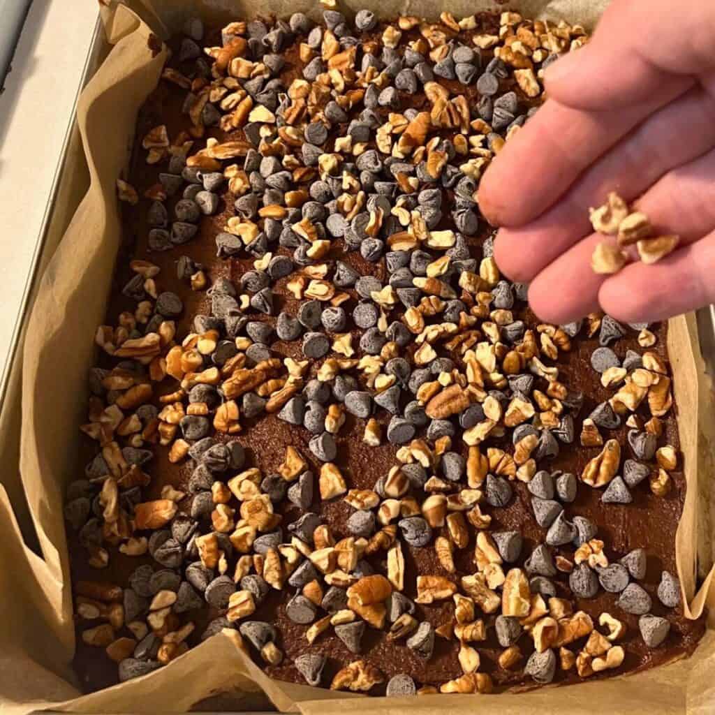 Woman sprinkling chopped pecans and chocolate chips to the top of a pan of brownie batter.