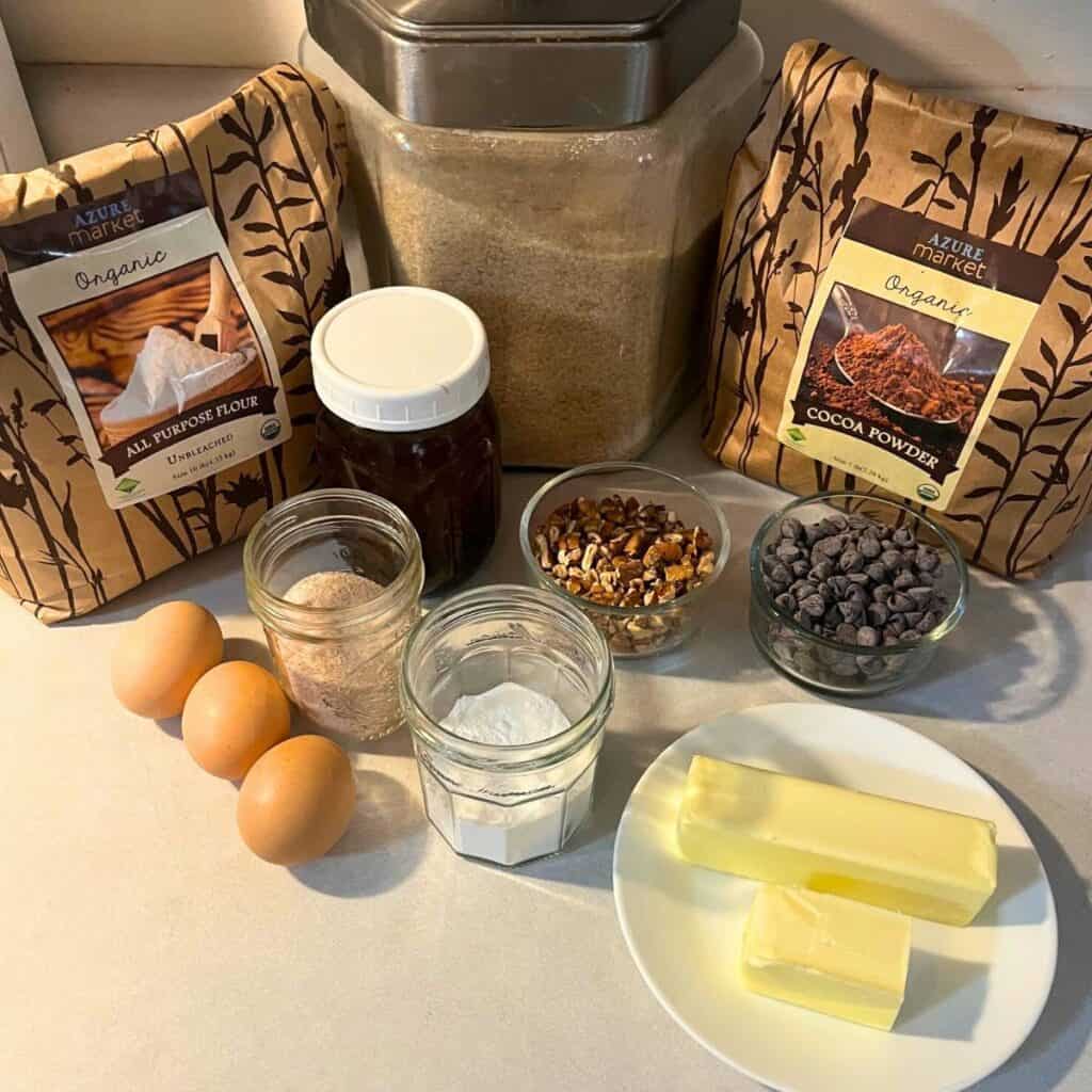 Ingredients for making double chocolate pecan brownies sitting on a kitchen counter. Pictured are all purpose flour, unsweetened cocoa powder, granulated sugar, vanilla extract, chopped pecans, chocolate chips, three eggs, salt, baking powder, and one and one half sticks of butter on a white plate.