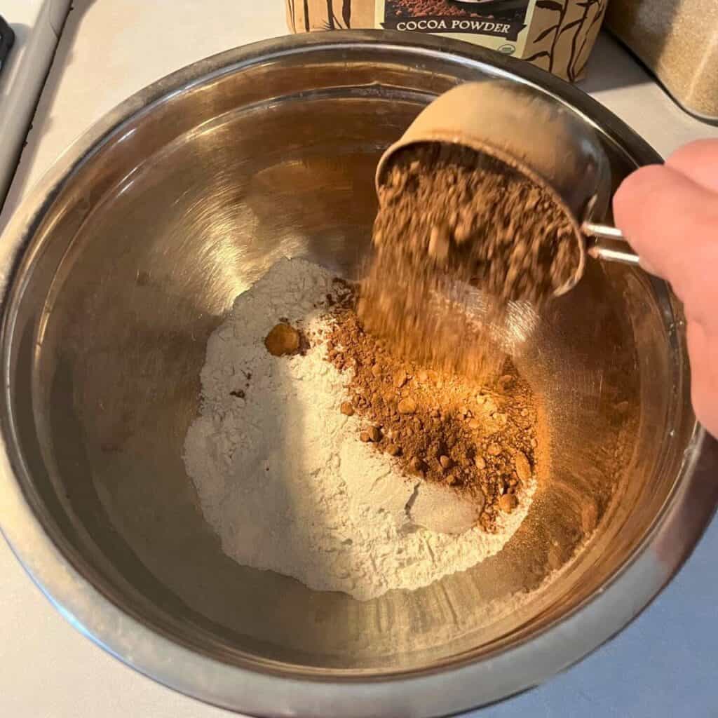Woman putting cocoa powder in a stainless steel mixing bowl with flour.
