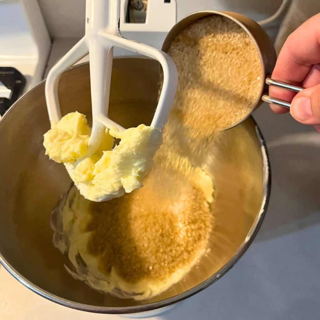Woman adding granulated sugar to a stand mixer with paddle attachment that has softened butter in it.