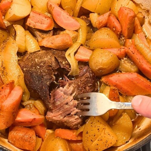 A beef pot roast cooked in an enameled cast iron Dutch oven with large diced potatoes, carrots, and onions. A silver fork is sticking out of the tender pot roast.