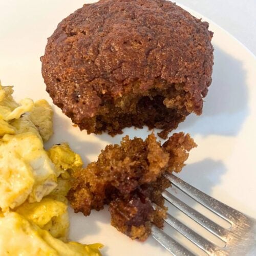 Woman cutting a cherry muffin on a white plate next to scrambled eggs.