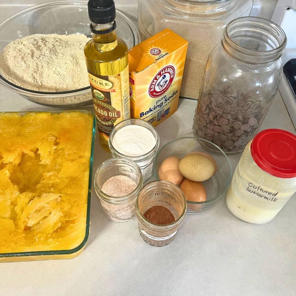 Ingredients for making chocolate chip pumpkin bread recipe sitting on a counter. Pictured are flour, pumpkin puree, avocado oil, salt, baking soda, baking powder, sugar, eggs, pumpkin pie spice, buttermilk, and chocolate chips.