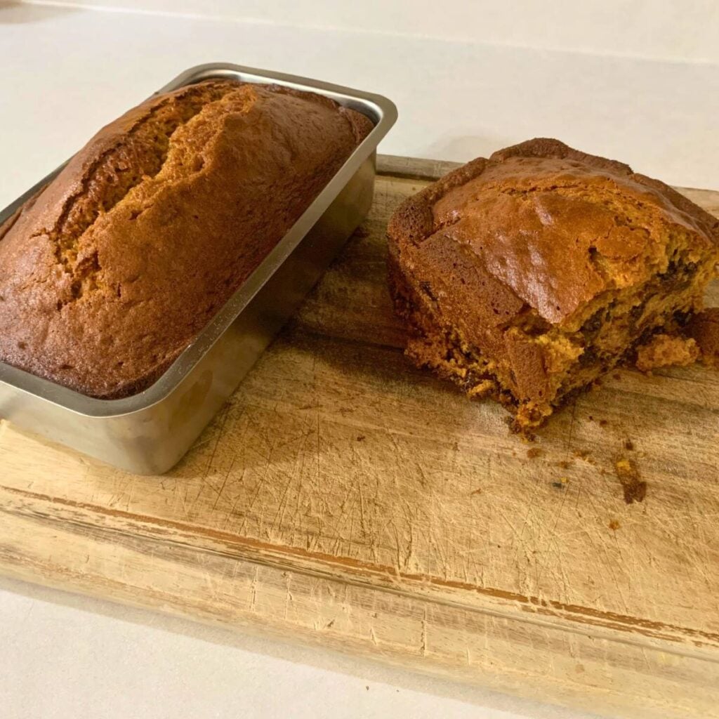 A loaf of cooked chocolate chip pumpkin bread in a stainless steel loaf pan alongside a half loaf on a wood cutting board.