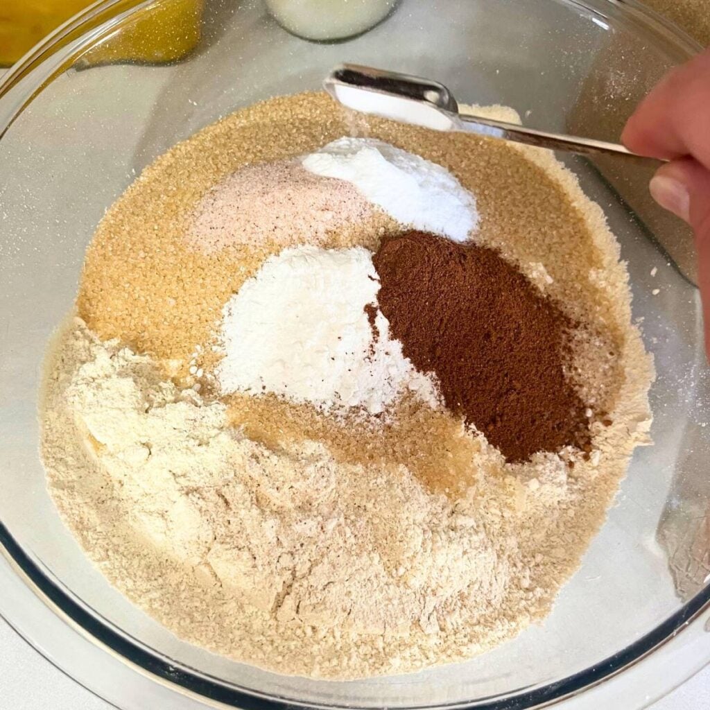 Woman adding baking soda to a glass mixing bowl that has freshly milled flour, sugar, baking powder, salt, and pumpkin pie spice inside.