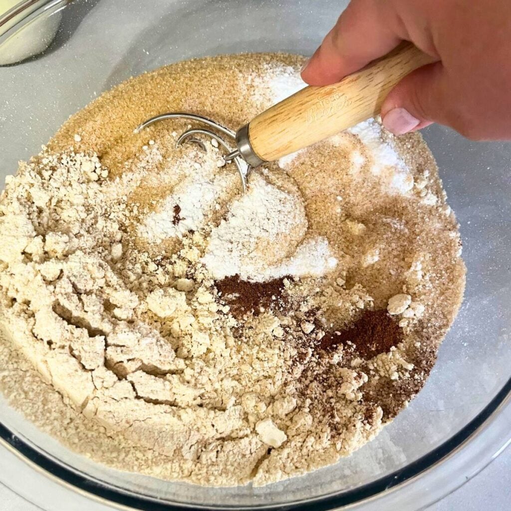 Woman stirring dry ingredients in a large glass mixing bowl using a Danish dough whisk.
