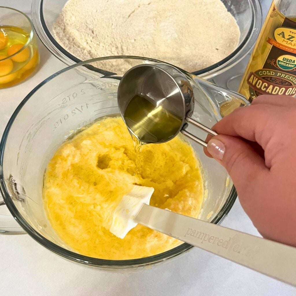 Woman adding cooking oil to a large mixing bowl that has eggs, milk, and pumpkin puree.