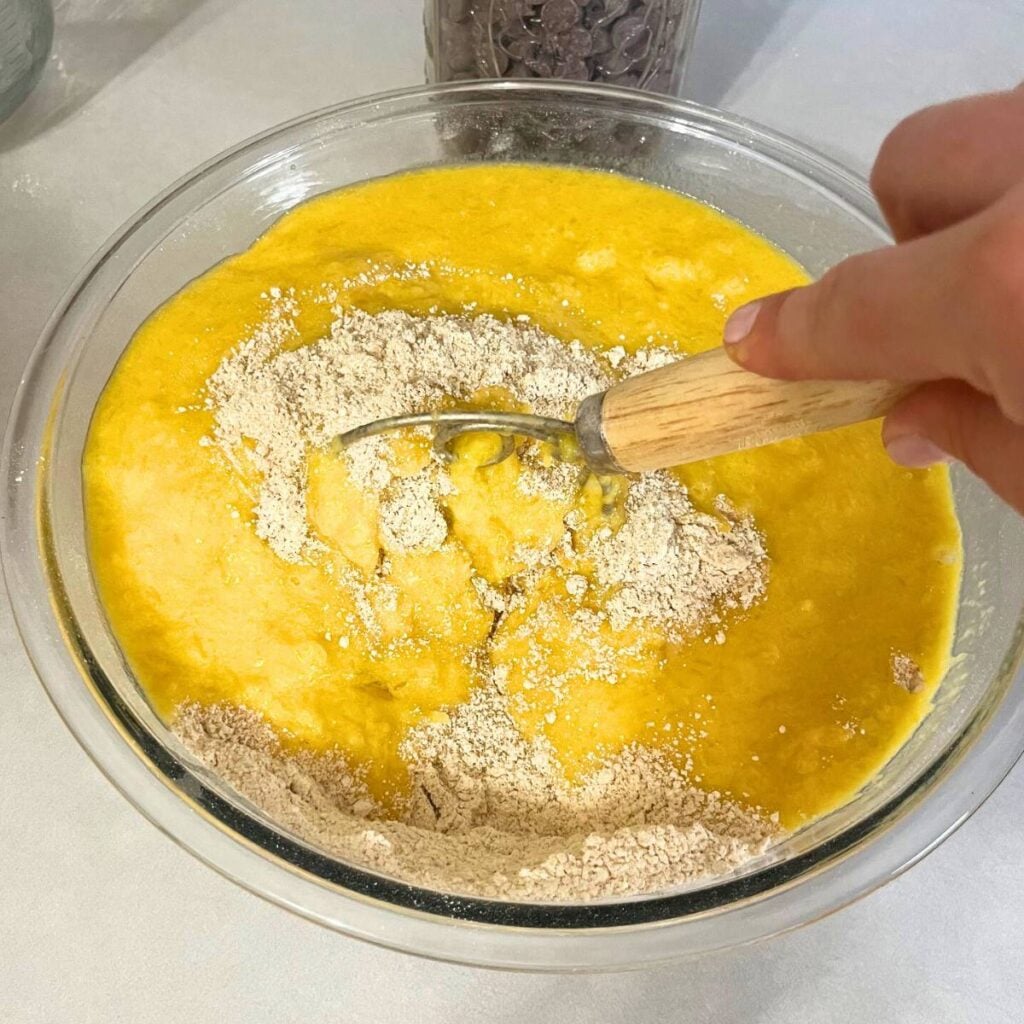 Woman mixing together wet ingredients into dry ingredients in a large glass bowl using a dough whisk.