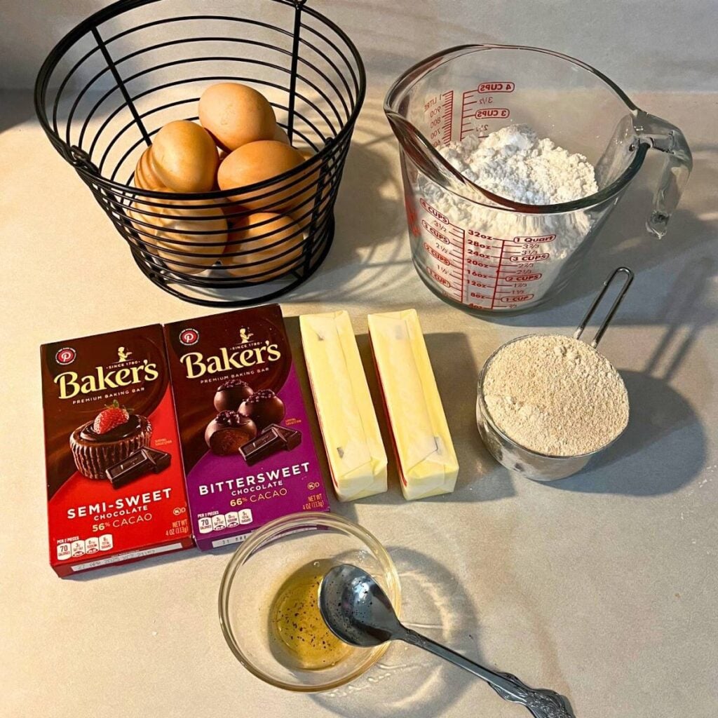 Ingredients for making chocolate lava cake sitting on a kitchen counter. Pictured from left to right and back to front are a basket of eggs, a measuring cup of powdered sugar, bittersweet and semisweet chocolate bars, two sticks of unsalted butter, a cup of wheat berries, and a small bowl of vanilla extract with a spoon.