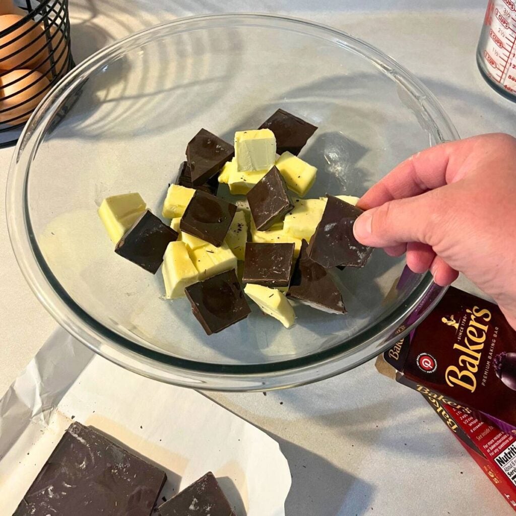 Woman adding chunks of bittersweet chocolate, semisweet chocolate, and tablespoons of unsalted butter to a large glass mixing bowl.