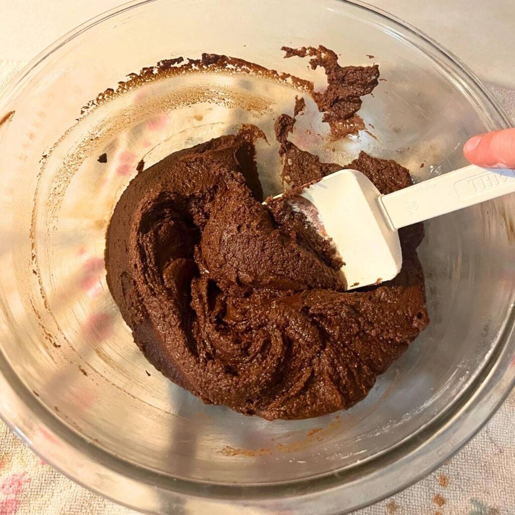 Woman stirring powdered sugar into melted butter and chocolate in a glass bowl using a white spatula.