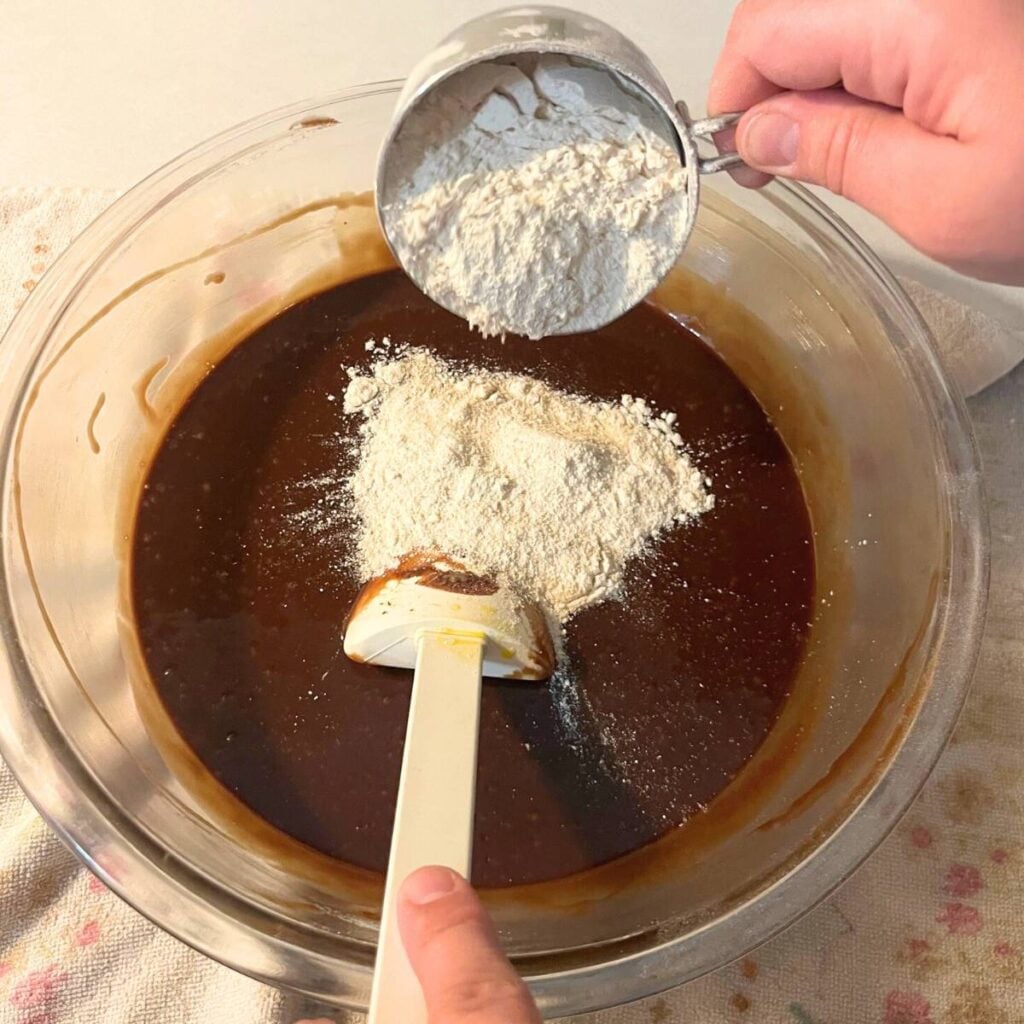Woman adding flour to chocolate lava cake batter in a glass bowl using a white spatula.