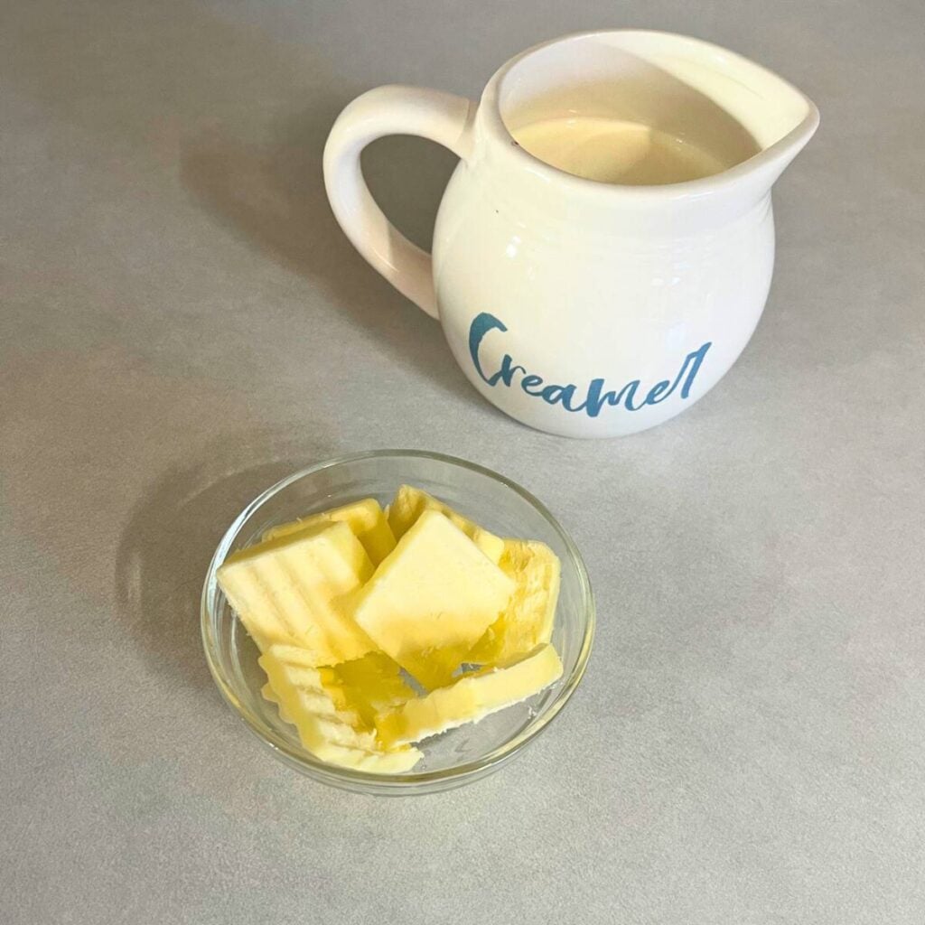 A small, white pitcher of heavy cream sitting on a counter next to a glass bowl of crinkle cut butter slabs.