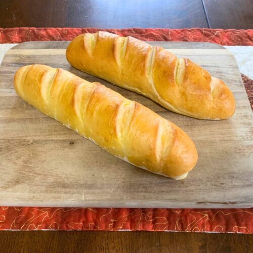 Two loaves of golden brown vegan French bread sitting on a wood cutting board on a table.
