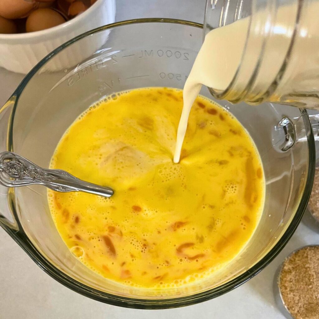 Woman pouring milk into a glass mixing bowl with eggs, cream, and vanilla extract inside.