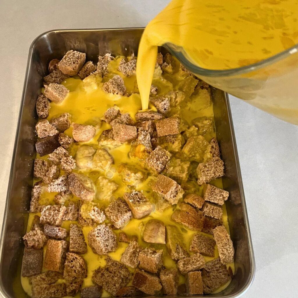 Woman pouring an egg mixture over cubes of bread in a 9x13 baking pan.