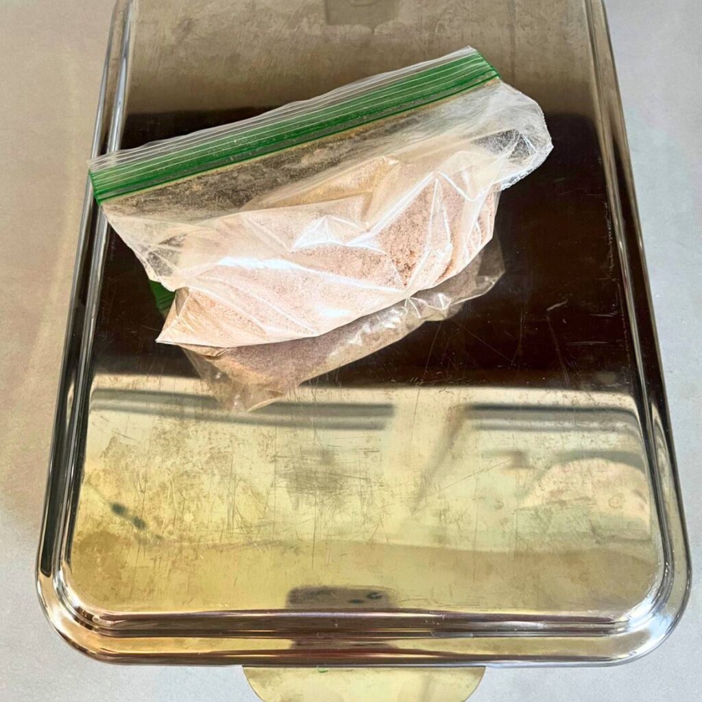 A stainless steel 9x13 pan with lid sitting on a counter. A sandwich bag of sweet topping sits on top of it.