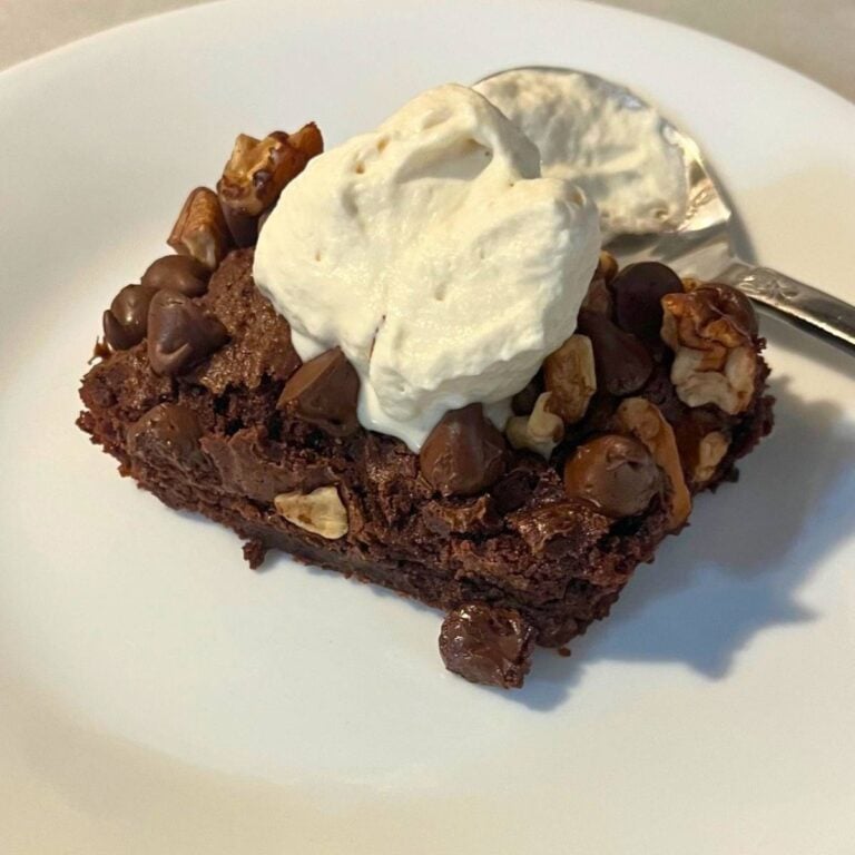 A chocolate brownie with nuts and whipped cream on top, sitting on a white plate next to a silver spoon.